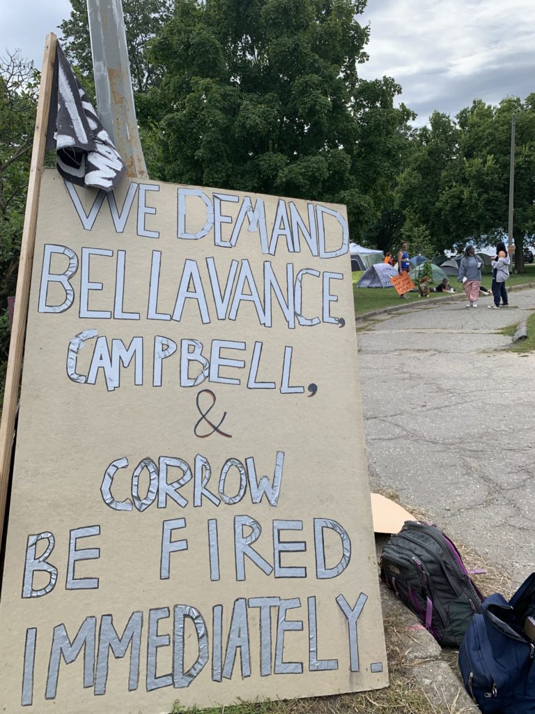 Cardboard sign with "WE DEMAND BELLAVANCE, CAMPBELL, & CORROW BE FIRED IMMEDIATELY" written in silver block letters outlined in black.